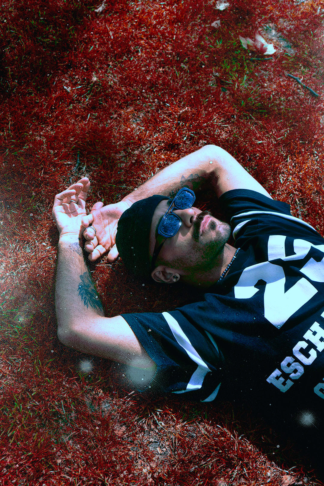 Astrologer and tarot reader Chris Corsini laying in the grass looking up into the distance, wearing a black beanie, black sunglasses and a black t-shirt,  offers horoscope predictions and astrology workshops online. 