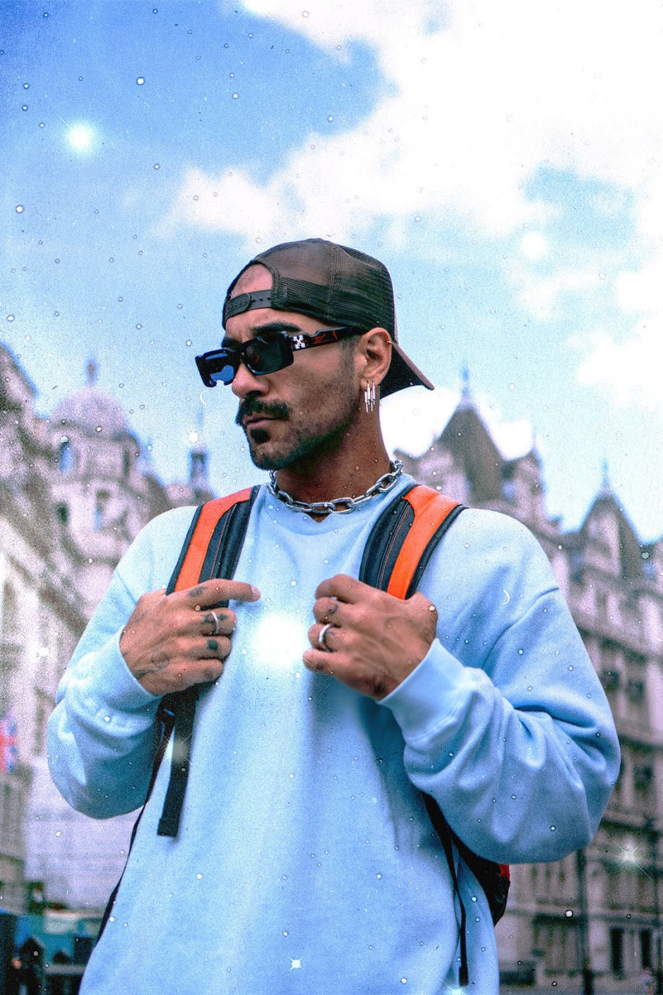 Astrologer and tarot reader Chris Corsini wearing a backwards black cap, large rectangle framed sunglasses, a silver chain necklace, a baby blue sweater and an orange backpack, looking off into the distance, offers horoscope predictions and astrology workshops online.